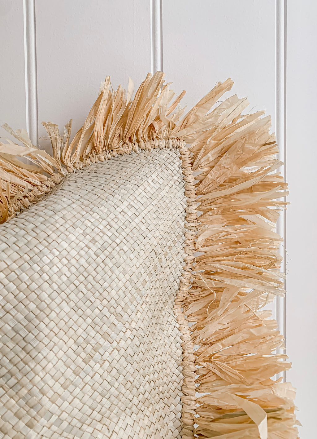 A Zali | Raffia cushion with a raffia fringe hanging on a white wall as home decor by Barre Living.