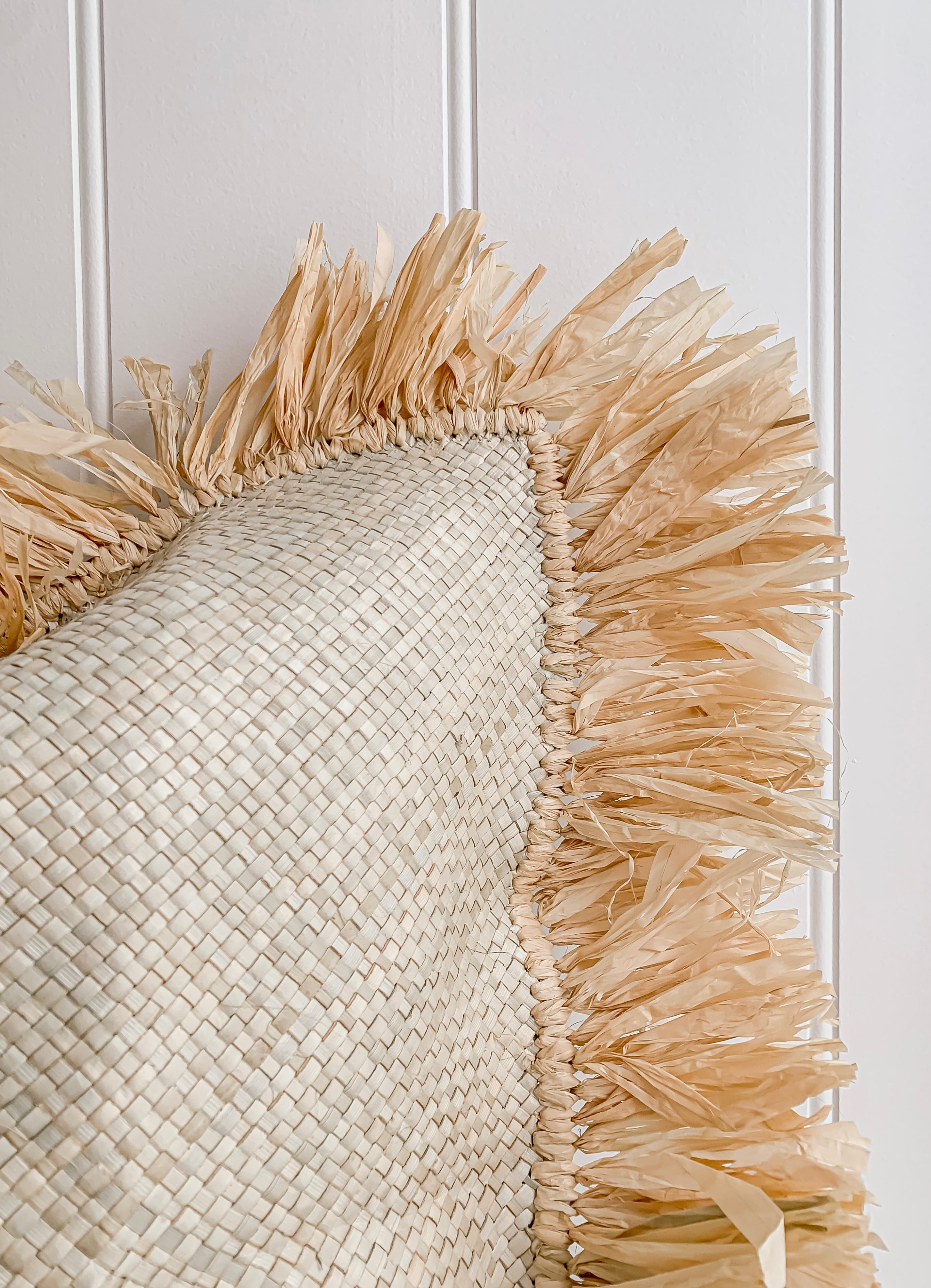 A Zali | Raffia cushion with a raffia fringe hanging on a white wall as home decor by Barre Living.